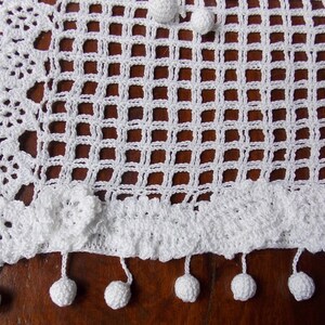 May include: White crocheted table runner with a square pattern and a scalloped edge. The runner has a fringe with white pom-poms.