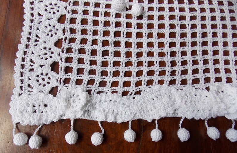 May include: White crocheted table runner with a square pattern and a scalloped edge. The runner has a fringe with white pom-poms.