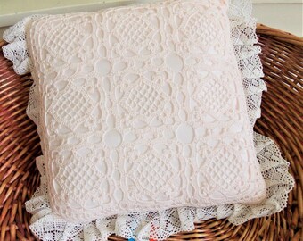 May include: A pink crocheted pillow with a white lace trim. The pillow is sitting in a wicker basket. The tag on the pillow reads "Collection Tricoteur d'Art Made in France."