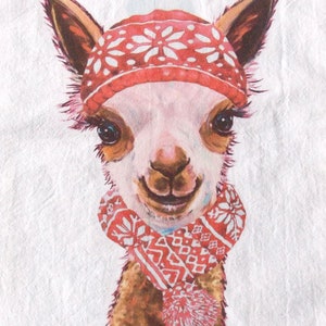 May include: A watercolor illustration of a smiling llama wearing a red hat and scarf with white snowflake patterns. The llama has brown fur, large eyes, and a pink nose. The artist's signature is in the bottom left corner.