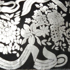 Peut inclure: Gros plan sur un tissu noir avec un motif floral blanc. Le motif présente de grandes fleurs détaillées et des accents en forme de ruban, créant un aspect texturé et élégant. Le contraste du tissu fait ressortir le motif floral.