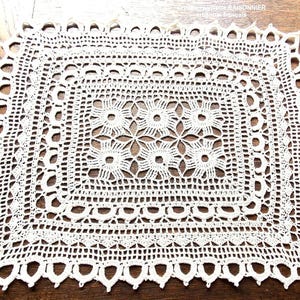 Puede incluir: Doily de crochet blanco con un diseño cuadrado en el centro. El doily tiene un borde festoneado.