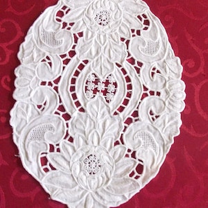 May include: An oval, white, embroidered doily with a floral design. The doily features detailed openwork and raised floral patterns, including roses and leaves. The background is a deep red fabric.