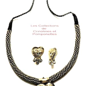 Könnte beinhalten: Eine goldfarbene Halskette mit einem herzförmigen Anhänger und passenden Ohrringen. Die Halskette besteht aus einer Kette mit einem schwarzen Band an jedem Ende. Der Anhänger und die Ohrringe sind mit klaren Strasssteinen verziert. Der Text "Les Collections de Crinolines et Pomponettes" ist im Bild sichtbar.
