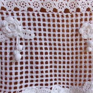 May include: White crocheted doily with a delicate floral design. The doily has a square shape with a grid pattern and two floral embellishments.