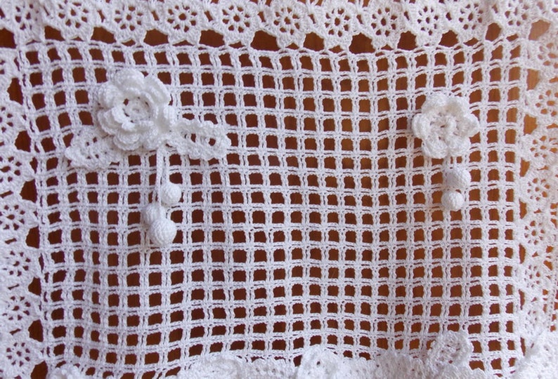 May include: White crocheted doily with a delicate floral design. The doily has a square shape with a grid pattern and two floral embellishments.