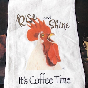 May include: White kitchen towel with a rooster illustration. The towel has the words "Rise and Shine" and "It's Coffee Time" printed on it. The rooster is depicted with red, orange, and white colors.