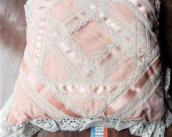 May include: A square, pink decorative pillow with a geometric design. The pillow features white lace trim and ribbon accents, creating a vintage aesthetic. The pillow has a ruffled lace edge.