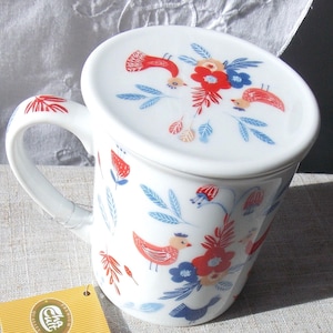 May include: White ceramic mug with a lid, featuring a floral and bird design in red, blue, and orange. The mug has a handle and the lid has a small knob.