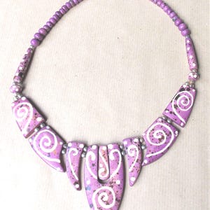 Peut inclure: Un collier composé d'une série de perles plates, géométriques, violettes et mouchetées, disposées en dégradé. Chaque perle est ornée de motifs en spirale blancs peints à la main. Le collier est complété par un rang de petites perles violettes.