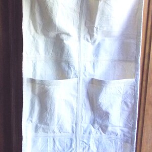 May include: Off-white fabric hanging organizer with multiple pockets. The organizer has a rectangular shape with a series of pockets sewn onto the front. It features a strap at the top for hanging. The fabric appears to be a light, possibly cotton, material.