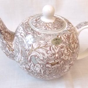 Puede incluir: Una tetera de cerámica blanca con un patrón floral marrón y gris. La tetera tiene una tapa con un pomo en la parte superior y un pico en el lateral. La tetera está sobre una superficie blanca.