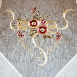 May include: A white tablecloth with embroidered floral design in shades of brown, gold, and red. The design features a bouquet of flowers with intricate swirls and loops.