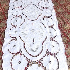 May include: White crocheted table runner with a delicate floral design. The runner features intricate cutouts and a scalloped edge.