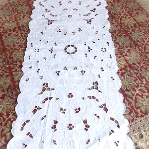 Peut inclure: Chemin de table blanc brodé avec un motif floral. Le chemin de table présente un motif répété de fleurs et de feuilles, avec des bords festonnés. La broderie détaillée crée un aspect délicat et élégant, adapté à une table à manger ou à une présentation décorative.