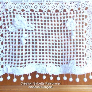 May include: White crocheted window valance with a delicate floral design and pom-pom trim. The valance is made with a grid pattern and has a scalloped bottom edge.  "Cr&eacute;ation Sylvette Raisonnier artisanat fran&ccedil;ais"