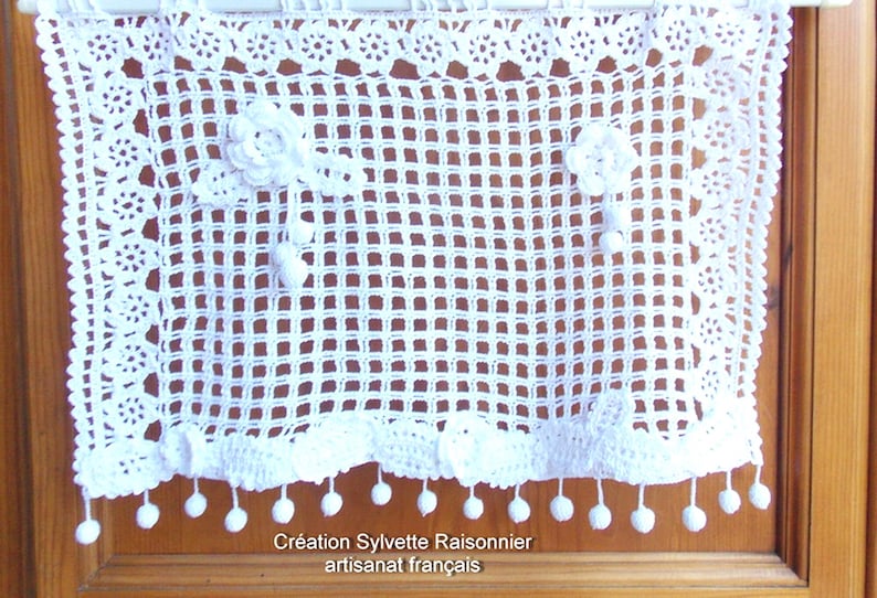 May include: White crocheted window valance with a delicate floral design and pom-pom trim. The valance is made with a grid pattern and has a scalloped bottom edge.  "Cr&eacute;ation Sylvette Raisonnier artisanat fran&ccedil;ais"
