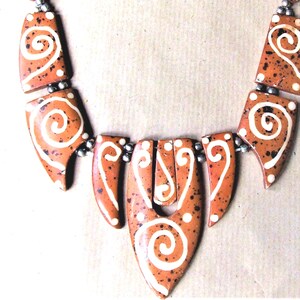 Peut inclure: Un collier composé de plusieurs segments en céramique de couleur terre cuite avec une finition mouchetée et des motifs en spirale peints à la main en blanc. Les segments sont reliés par de petites perles métalliques argentées.