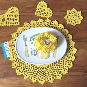 May include: A set of yellow crocheted doilies, including a large round doily, two heart-shaped doilies, and a smaller round doily. The doilies are placed on a wooden surface with a white plate in the center.