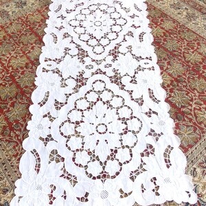 Peut inclure: Chemin de table en dentelle blanche avec un motif floral et géométrique. Le chemin de table présente des motifs découpés de fleurs et de cercles, créant un effet ajouré. Les bords sont festonnés. Le chemin de table est posé sur un tapis rouge et beige à motifs.
