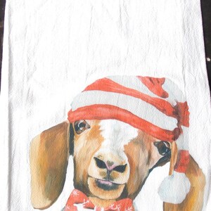 May include: White kitchen towel featuring a watercolor illustration of a goat wearing a red and white striped hat and a red bow tie with snowflakes. The goat has brown and white fur and is holding a small ornament.