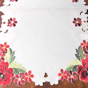 May include: White table runner with embroidered red flowers and green leaves. The runner has a scalloped edge and is decorated with a floral pattern. The flowers have yellow centers and are arranged in clusters.