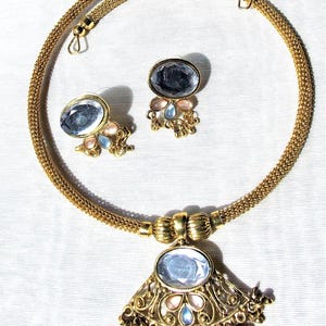 May include: A gold chain necklace with a pendant featuring a large oval-shaped clear stone surrounded by smaller pink and blue stones. The pendant is attached to a gold chain with a decorative design. The necklace comes with a pair of matching earrings with a similar design.