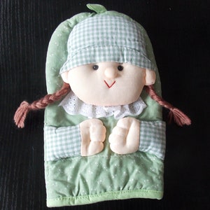 Puede incluir: Manopla de horno de tela verde y blanca a cuadros, con forma de niña con coletas marrones. La niña lleva un sombrero verde y blanco a cuadros y una cara sonriente.