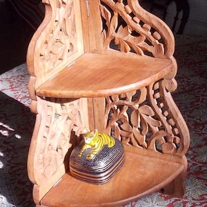 Peut inclure: Étagère d'angle en bois clair à deux niveaux, ornée de sculptures florales. Une petite boîte décorative avec une figurine de chat jaune et noir repose sur l'étagère inférieure. Le bois est de couleur marron clair.