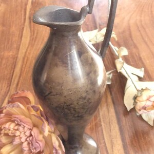 Könnte beinhalten: Eine bronzene Vase mit Henkel, rundem Korpus und einem ausgestellten Fuß. Die Vase steht auf einer Holzoberfläche.
