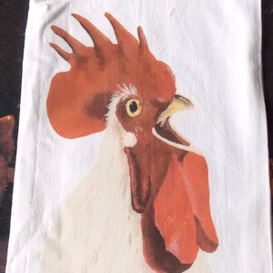 May include: White kitchen towel featuring a vibrant painting of a rooster's head. The rooster has a bright red comb and wattle, a yellow beak, and a detailed eye. The artist's signature, "Lee Keller," is visible.