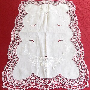 Op de afbeelding: Een witte, rechthoekige doily met een geschulpte rand en ingewikkelde kanten details. Het midden heeft geborduurde bloemmotieven en gebogen stelen. De doily ligt op een rode ondergrond.