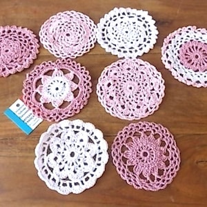 Könnte beinhalten: Eine Sammlung handgefertigter Häkeluntersetzer in verschiedenen Rosa- und Weißtönen. Die Untersetzer weisen komplizierte, spitzenartige Muster auf und sind auf einer Holzoberfläche angeordnet. Einige Untersetzer haben eine durchgehend rosa Mitte, andere eine weiße Mitte.