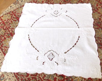 Puede incluir: Un mantel individual blanco, rectangular y bordado, con borde festoneado. El centro presenta un gran diseño de círculo abierto con calados florales y geométricos. El mantel individual está colocado sobre una alfombra roja y marrón estampada.