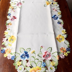 Op de afbeelding: Witte tafelrunner met geborduurd bloemenmotief. Het ontwerp bevat kleurrijke viooltjes en bladeren. De tekst "les collections de Crinolines et Pomponettes" is zichtbaar in de rechterbovenhoek.