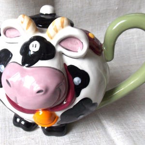 Op de afbeelding: Een witte theepot van keramiek in de vorm van een koe met zwarte vlekken. De theepot heeft een groene handgreep en een roze neus. De koe draagt een zwart-witte hoed en een rood-oranje halsband.