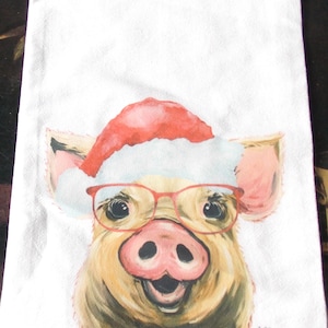 May include: White kitchen towel featuring a whimsical illustration of a pig wearing a Santa hat and red glasses. The pig has a pink snout and ears, and a smiling expression. The towel is made of a soft, absorbent material.