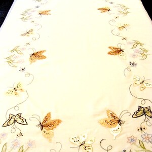 Peut inclure: Chemin de table brodé de couleur crème avec un motif de papillons. Les papillons sont dans des tons de marron, crème et blanc, avec de délicats accents floraux et de feuilles. Le texte "collectors of Crinolines on Pompadours" est dans le coin supérieur gauche.