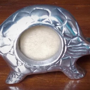 Può includere: Un portafoto a forma di tartaruga argentato con un'apertura rotonda per una foto. Il telaio è in metallo e ha una superficie testurizzata.