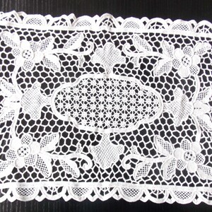 May include: A white, rectangular doily with an intricate lace design. The doily features floral and geometric patterns, including flowers, leaves, and a central oval shape. The edges are scalloped, adding a delicate touch to the handmade textile.