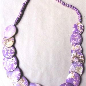 Peut inclure: Un collier composé d'une série de grandes perles plates et circulaires arborant un motif moucheté violet et crème. Le collier se termine par des perles rondes violettes plus petites vers le haut, avec un fermoir métallique simple.