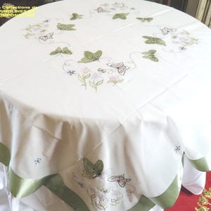 May include: A round white tablecloth with embroidered butterflies and floral designs. The butterflies are in shades of green and pink, with delicate floral accents. The edge of the tablecloth has a green satin trim. The text "Les Collections de Crinolines Pomponnettes" is in the upper left corner.