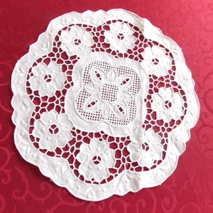 Puede incluir: Un tapete blanco circular con un intrincado diseño floral. El tapete presenta un motivo floral central rodeado por un anillo de flores y un borde festoneado. El diseño calado crea una apariencia delicada y elegante, adecuada para la decoración del hogar.