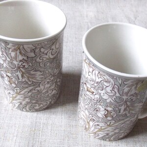 Op de afbeelding: Twee witte keramische mokken met een bloemenpatroon in grijstinten en bruin. De mokken hebben een handvat en zijn perfect om van een warme drank te genieten.