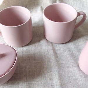 May include: A pale pink ceramic tea set comprising two handled mugs, a lidded sugar bowl, and a creamer jug. The items are arranged on a light-coloured, textured surface.