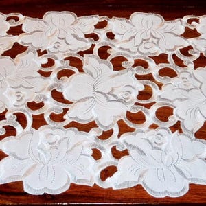 Peut inclure: Un chemin de table en dentelle blanche avec un motif floral. Le chemin de table est fait d'une dentelle délicate et complexe et présente un motif de fleurs répétitif.