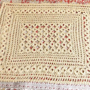 May include: A cream-coloured, rectangular crochet doily with an intricate openwork design. The doily features a repeating pattern of circles and open spaces, creating a delicate and textured appearance. The edges are finished with a scalloped border.