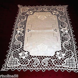 May include: A rectangular, off-white lace doily with a central panel and a decorative, openwork border. The border features floral and geometric patterns. The doily is placed on a dark red surface.