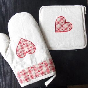 Puede incluir: Un juego de dos agarraderas de cocina beige con diseños de corazones rojos. Las agarraderas están hechas de un tejido suave y texturizado y presentan un patrón de cuadros rojos y blancos.