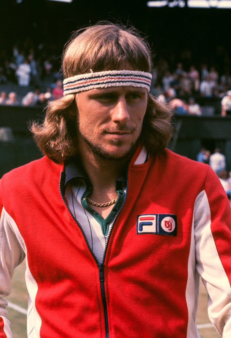 May include: A portrait of a man wearing a red and white track jacket with a FILA logo. He has long, light brown hair and a white headband with red and blue stripes. A gold chain is visible at his neck.